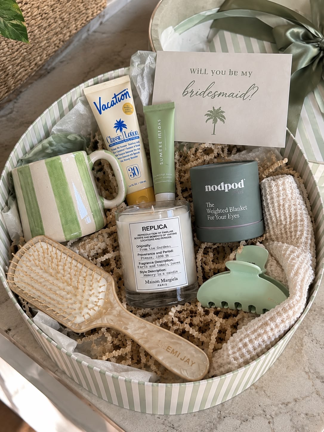 An open round bridesmaid box with a candle, striped mug, hair brush and claw clip nestled in paper straw
